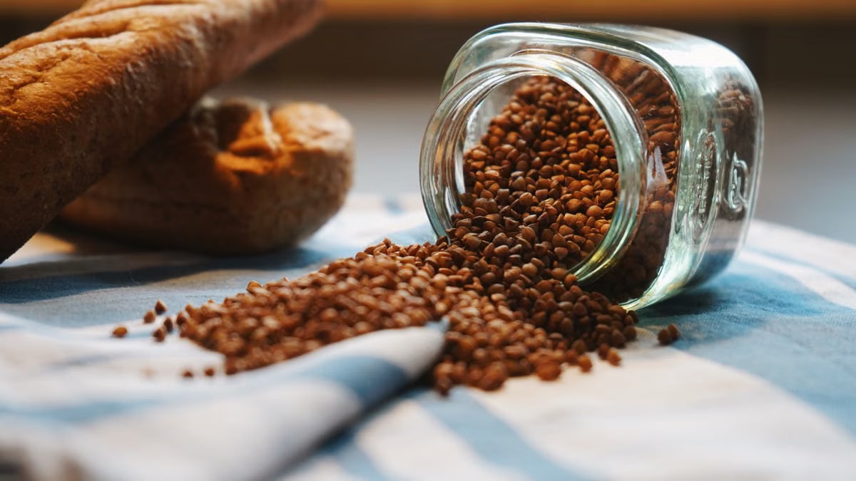 A rustic scene with buckwheat spilling from a jar next to fresh bread on a checkered cloth.